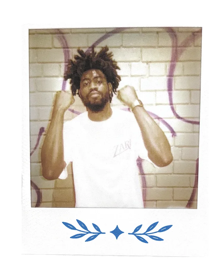 A member against a graffitied wall, hands up, in the Zari tee.